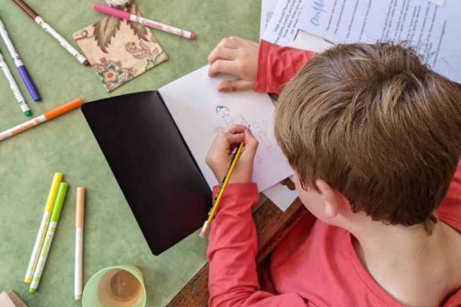 Boy drawing in a sketchbook, there are pens scattered on the table.