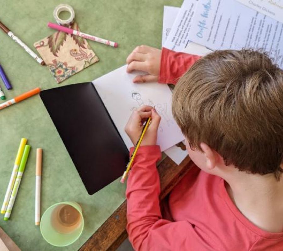 Boy drawing in a sketchbook, there are pens scattered on the table.