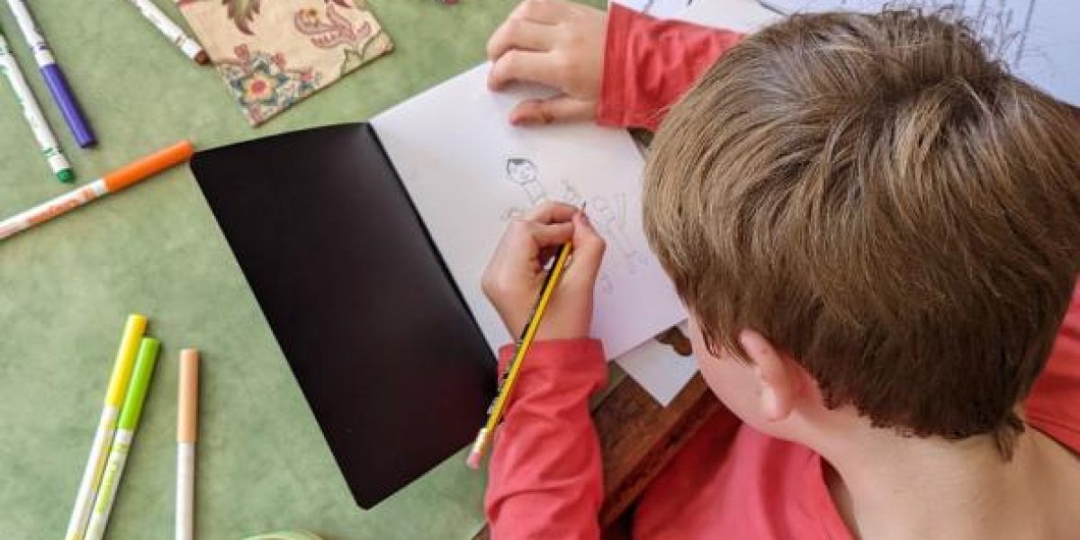 Boy drawing in a sketchbook, there are pens scattered on the table.