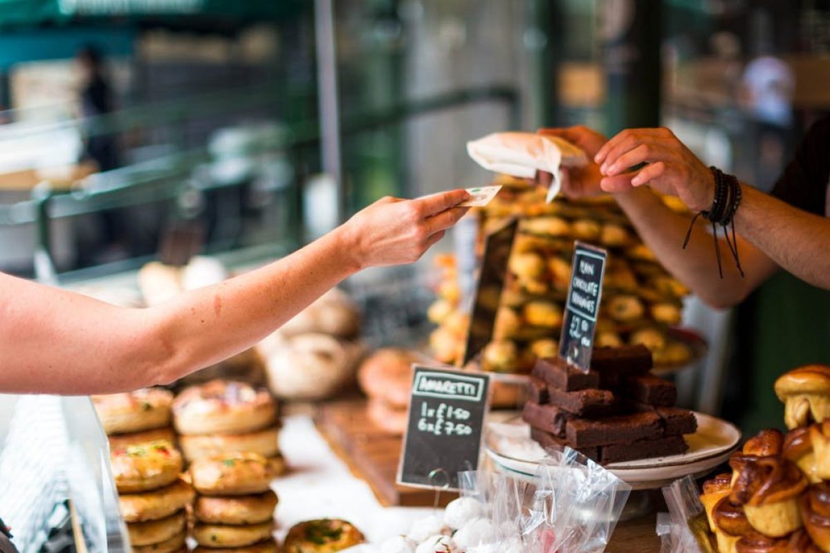 Close up color image depicting a cash transaction between a customer and the owner of a bakery stall at an outdoor food market in London, UK. The customer is paying using a five pound note for what appears to be some kind of cake or pastry. In the background the contents of the market stall - cakes, brownies, sweet pastries - are blurred out of focus. Room for copy space.