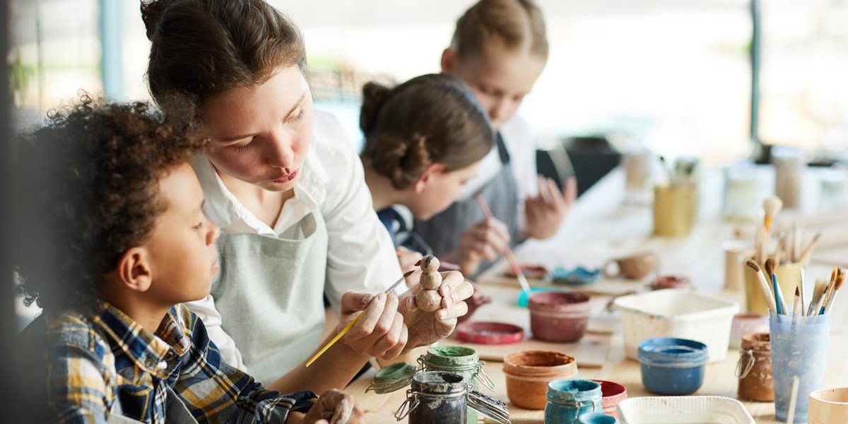 Young teacher showing one of pupils clay item and giving advice about painting it