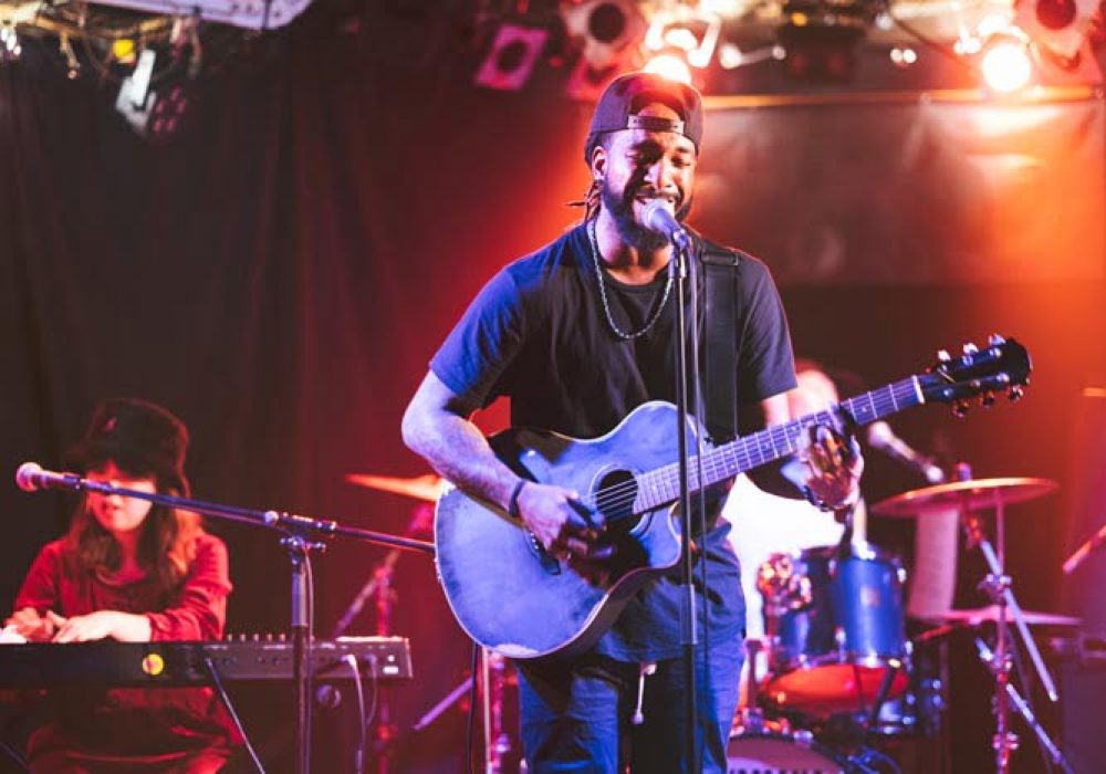 A black male guitarist is singing and playing the acoustic guitar on stage.