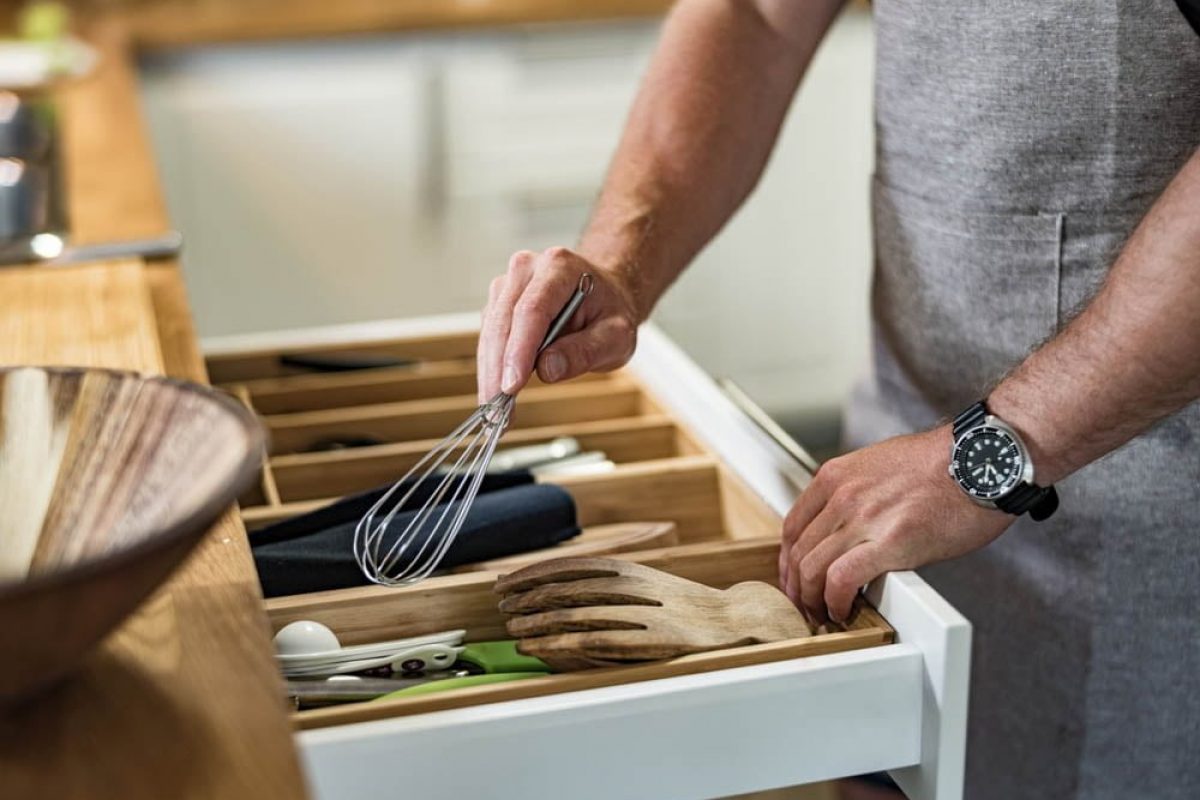 Hands of Young Caucasian man taking the wire whisk from the drawer  in home kitchen. He is wearing casual clothes with chef's apron. He is preparing Italian inspired meal with fresh pasta, meat balls and tossed salad with fresh vegetables and vinaigrette.