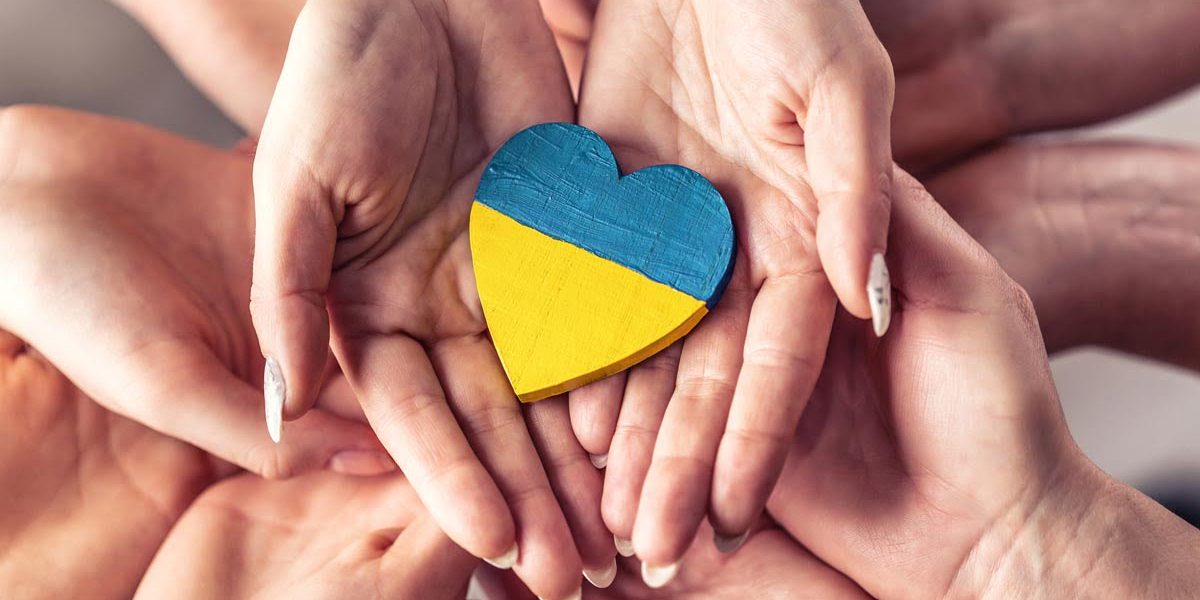 We stand with Ukraine symbol as numerous hands hold one another in unity with a wooden heart painted in Ukrainian flag colors on top.