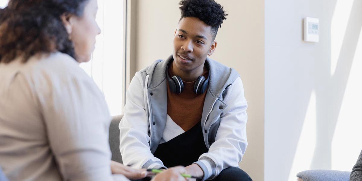 In therapy, the mid adult female counselor talks to the attentive young man and his unseen sister.