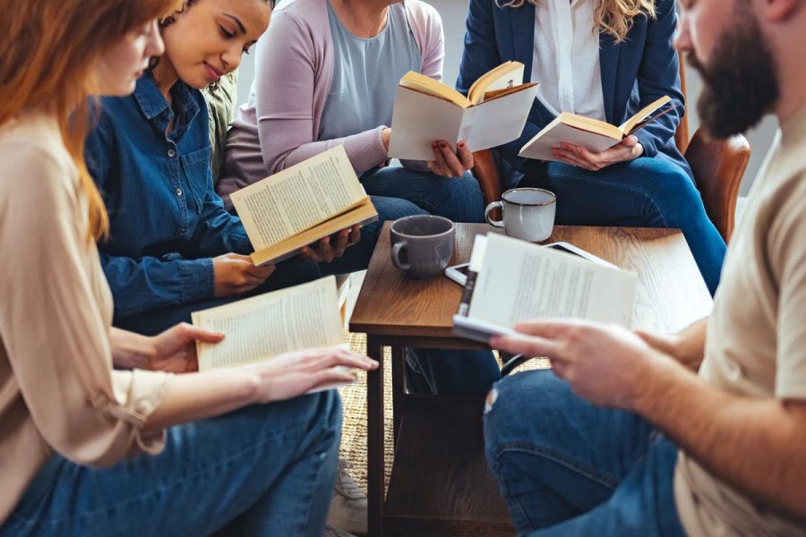 Small group of people with a mixed age range sitting at a table, discussing and reading books together.
