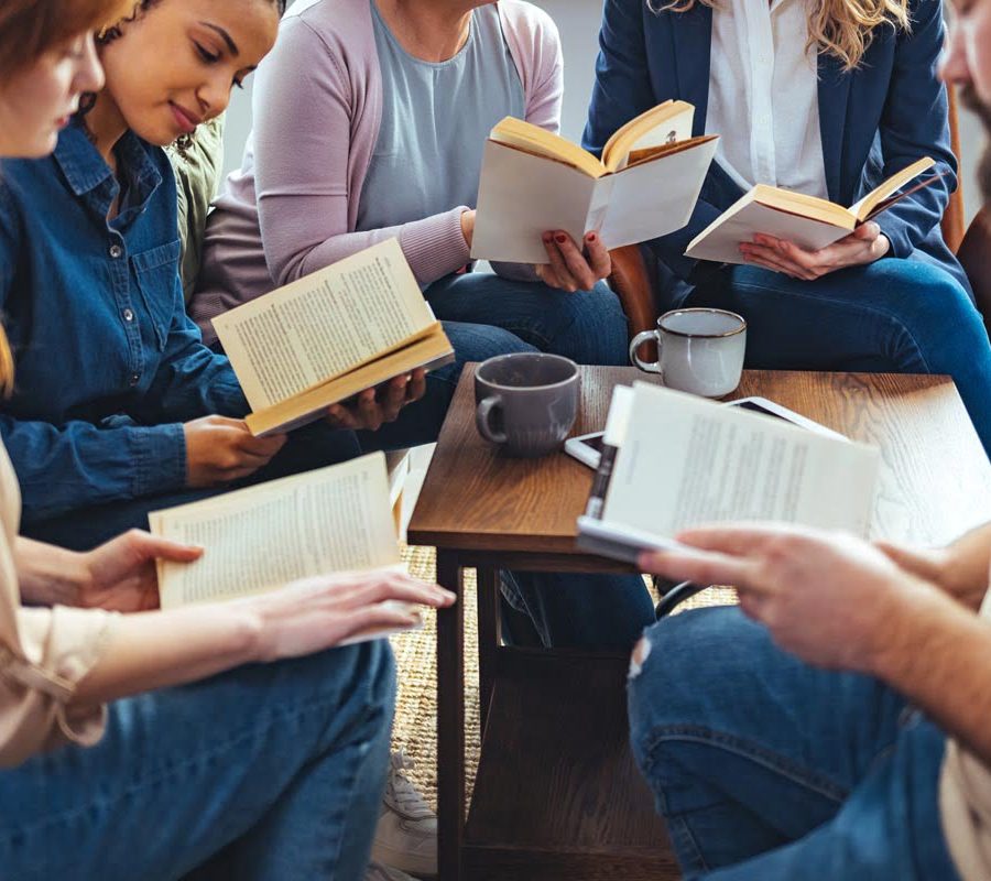 Small group of people with a mixed age range sitting at a table, discussing and reading books together.