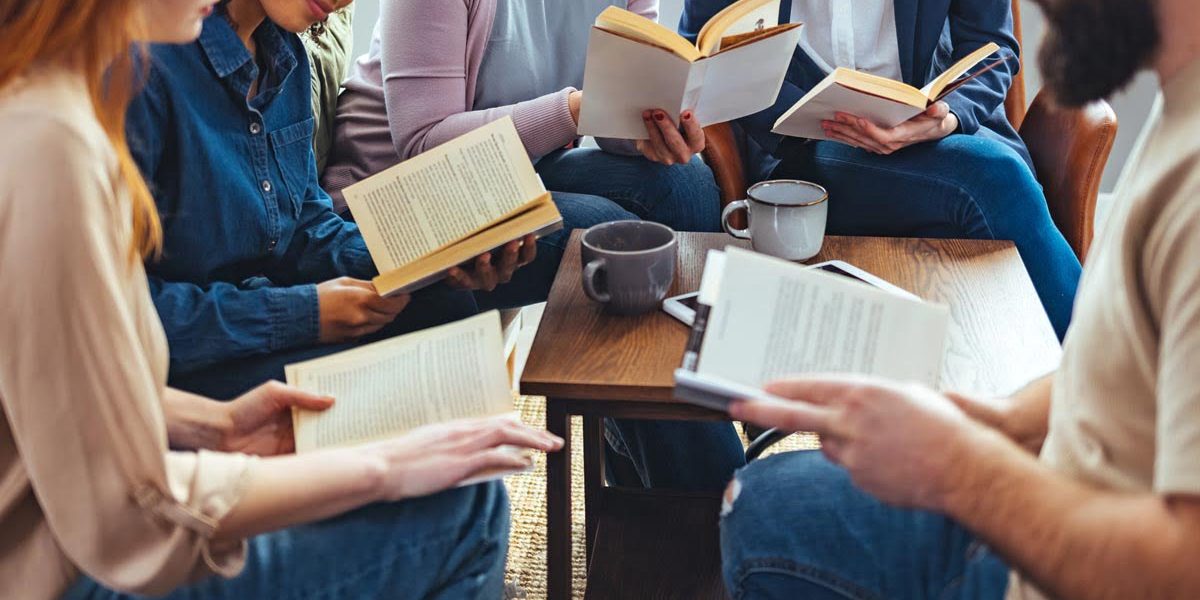 Small group of people with a mixed age range sitting at a table, discussing and reading books together.
