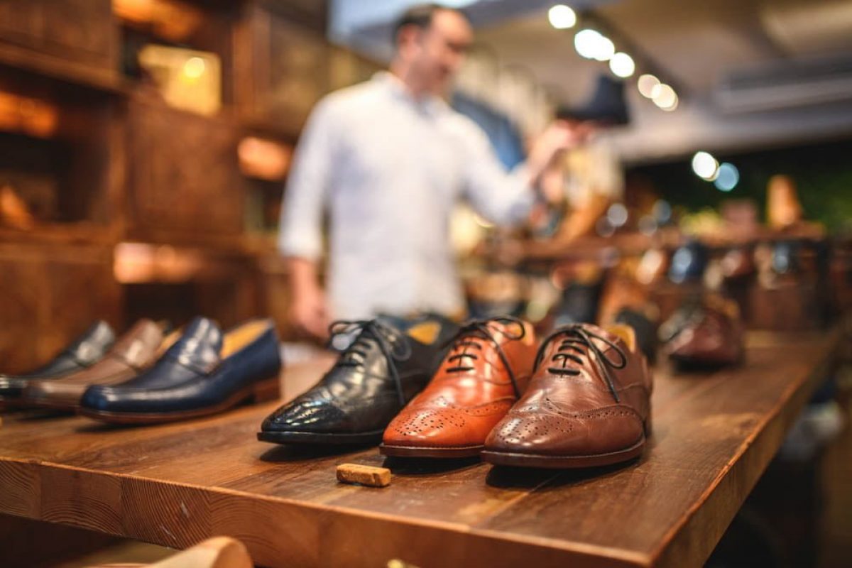Close-up shot of elegant men's brogue shoes in a luxury leather footwear store. The shoes are displayed in a row on a wooden shelf. Defocused male customer in the background.