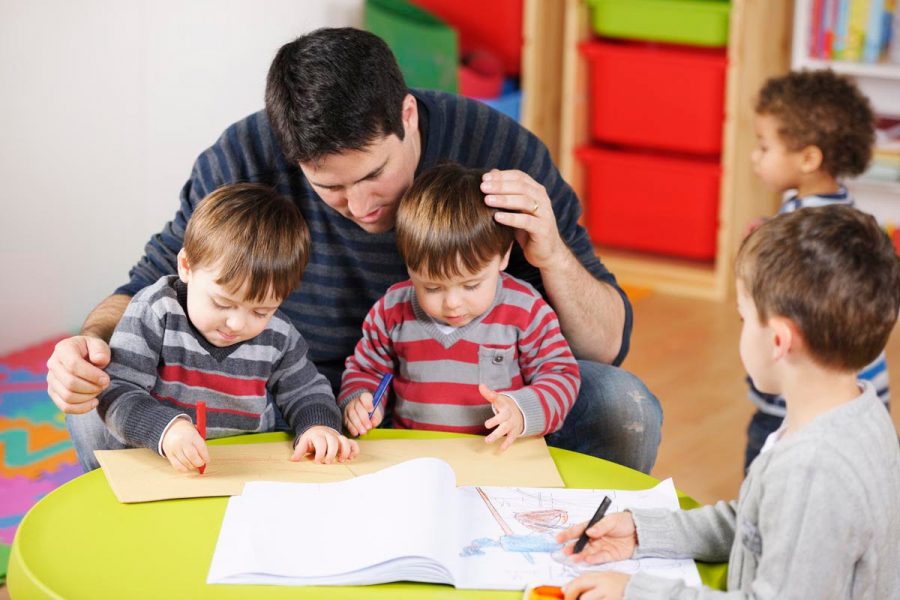 Carer/ childminder/teacher supervising group of toddlers and litle boy doing artwork in a nursery setting.