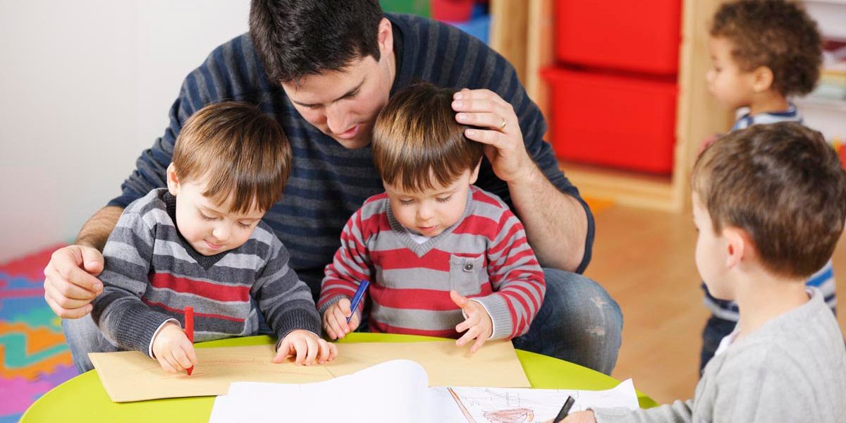 Carer/ childminder/teacher supervising group of toddlers and litle boy doing artwork in a nursery setting.