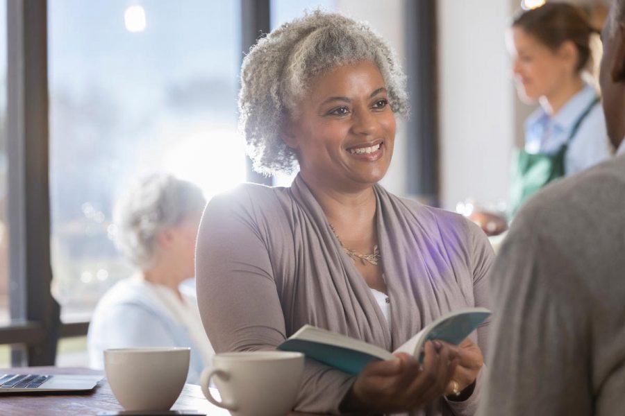 Smiling senior woman discusses a book with her unrecognizable husband while having coffee in local coffee shop.