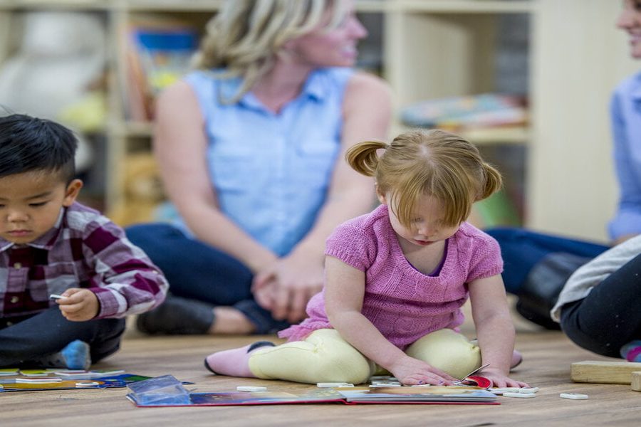 A group of kids and their mothers are indoors in a preschool. The kids are playing with toys and books in the foreground while their mothers chat in the background.