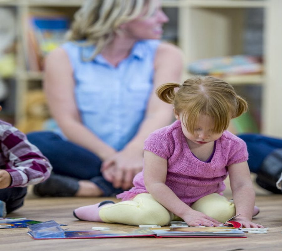 A group of kids and their mothers are indoors in a preschool. The kids are playing with toys and books in the foreground while their mothers chat in the background.