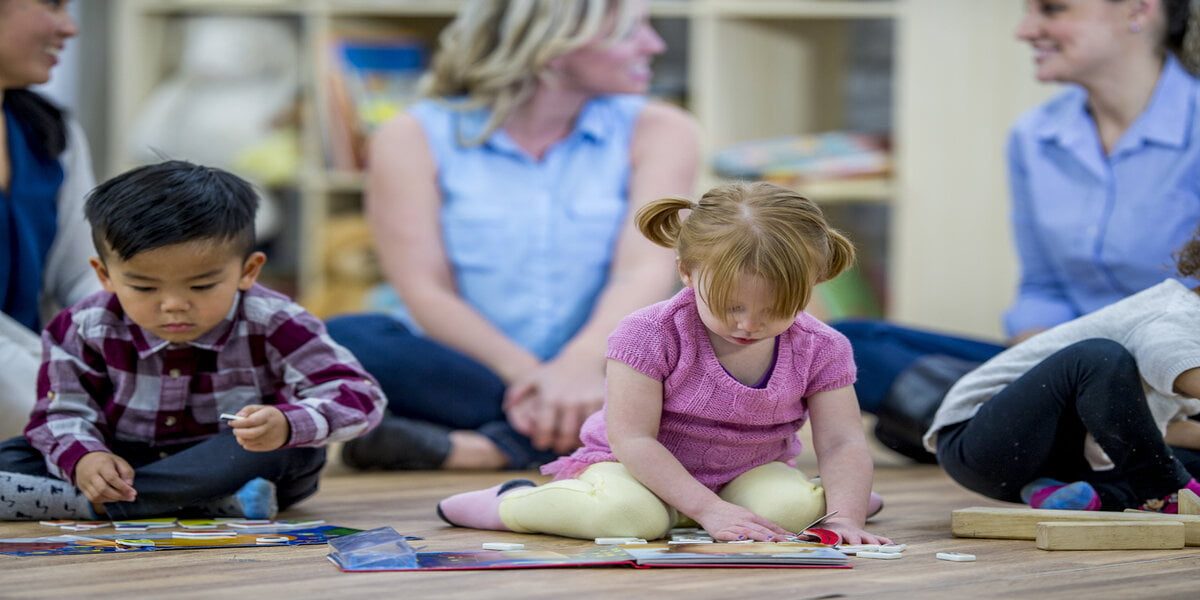 A group of kids and their mothers are indoors in a preschool. The kids are playing with toys and books in the foreground while their mothers chat in the background.