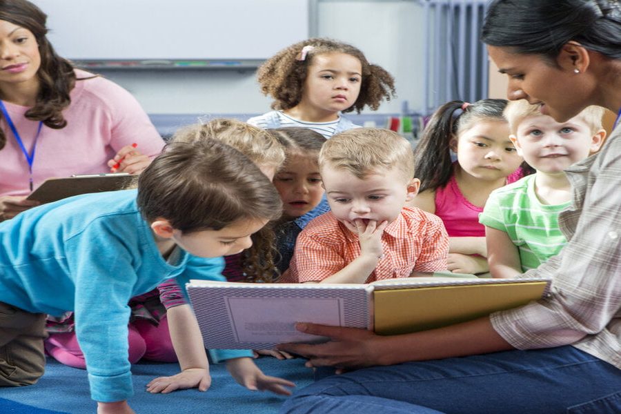 Female teacher giving a lesson to nursery students. They are sitting on the floor and there is a teacher taking notes.