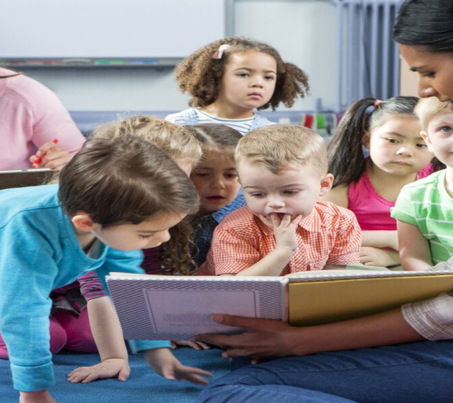 Female teacher giving a lesson to nursery students. They are sitting on the floor and there is a teacher taking notes.